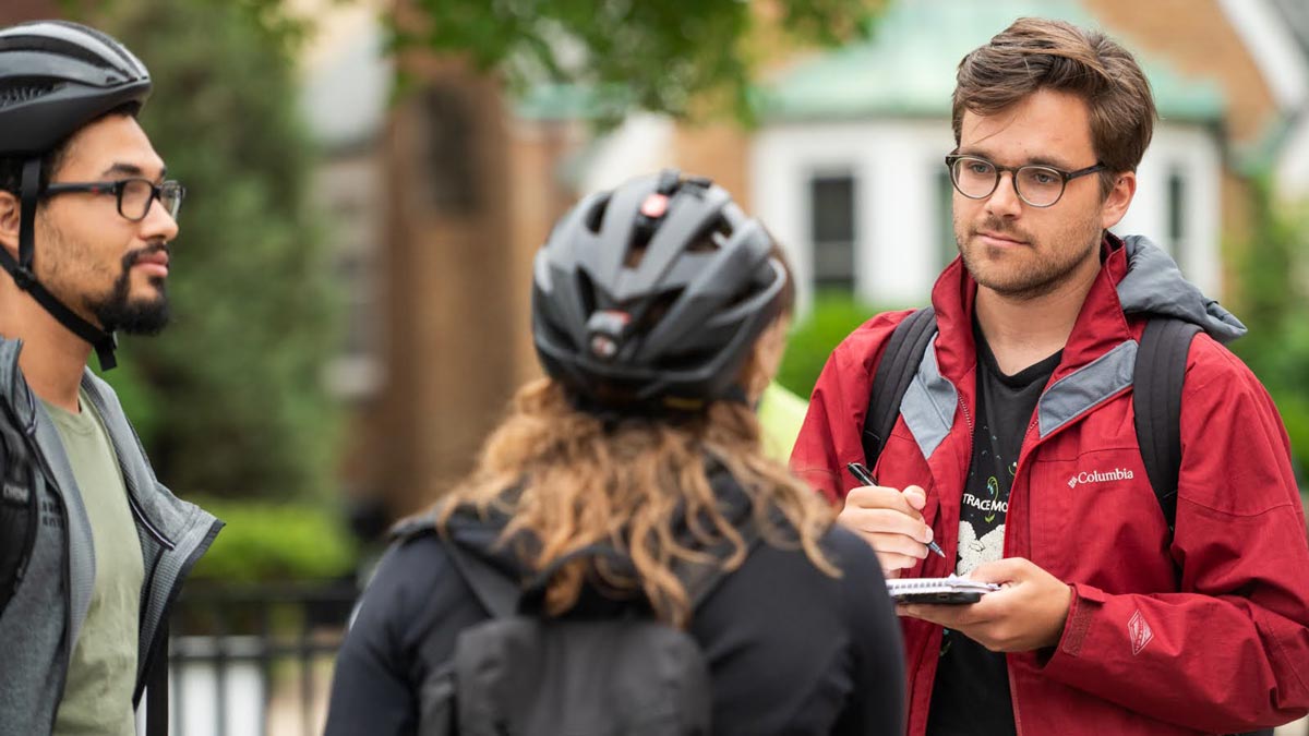 Three people talking outside, one taking notes with a notepad.