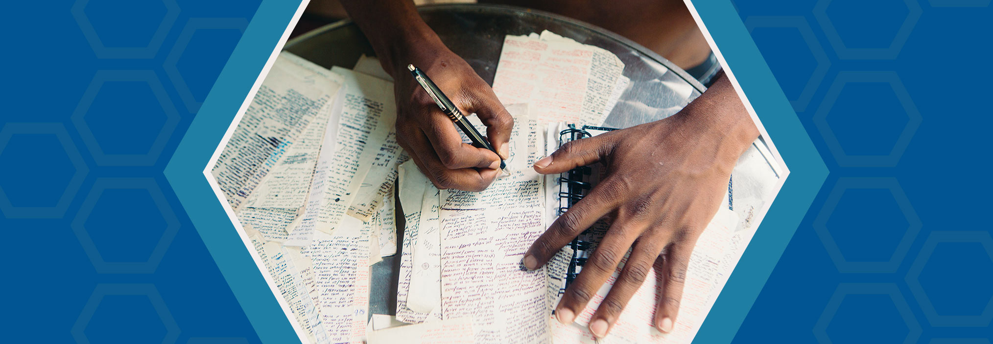 Hands writing on a notebook surrounded by handwritten papers, on a blue background with hexagon shapes.
