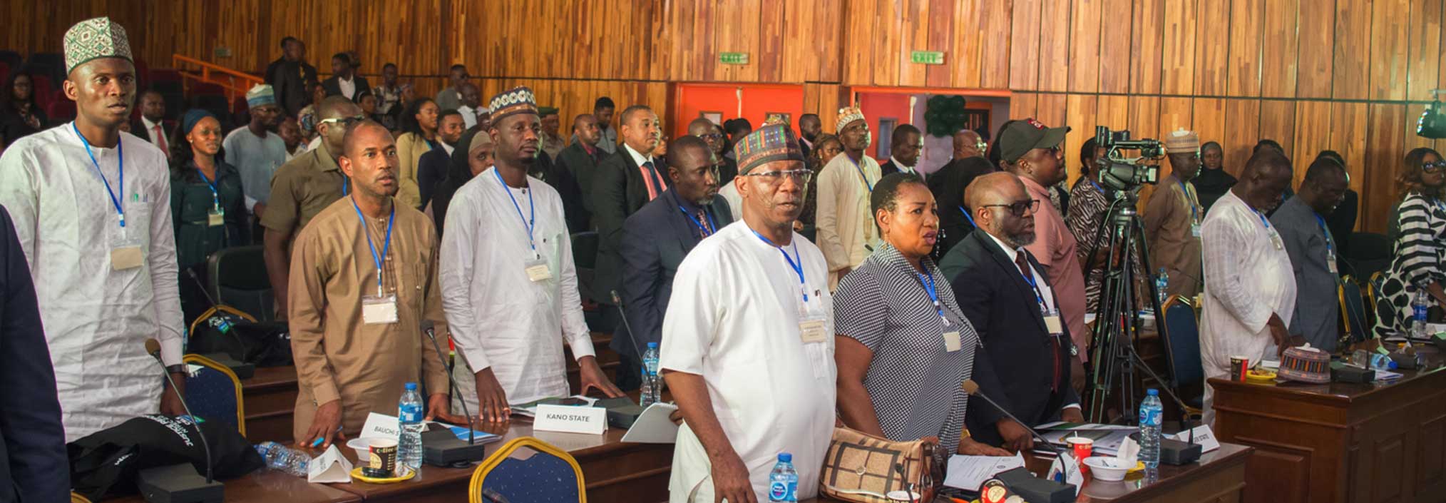 A group of people standing in a conference room with wooden walls, wearing lanyards and name tags.